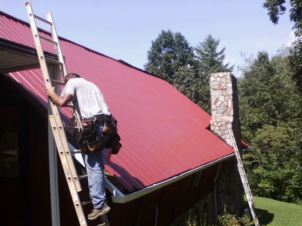 Roofer on a ladder beside a red metal roof and stone chimney — Soffit & Fascia Repair by Roofing Brothers in Othello